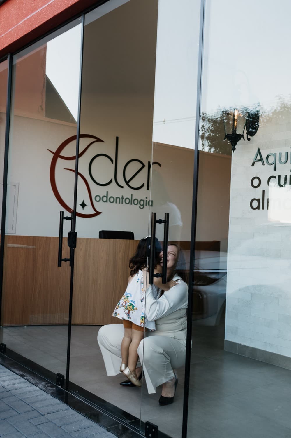 Fachada de vidro da Clínica Cler com a Dra. Jessica acolhendo uma criança na entrada
